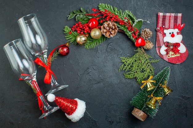 Vista anterior del estado de ánimo navideño con copas de cristal caído ramas de abeto calcetín de árbol de Navidad sombrero de santa claus sobre fondo oscuro