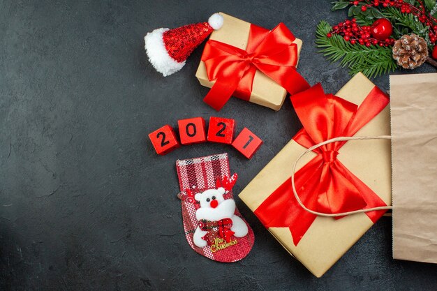 Vista anterior del estado de ánimo navideño con hermosos regalos con cinta roja y números calcetín de Navidad sombrero de santa claus sobre fondo oscuro