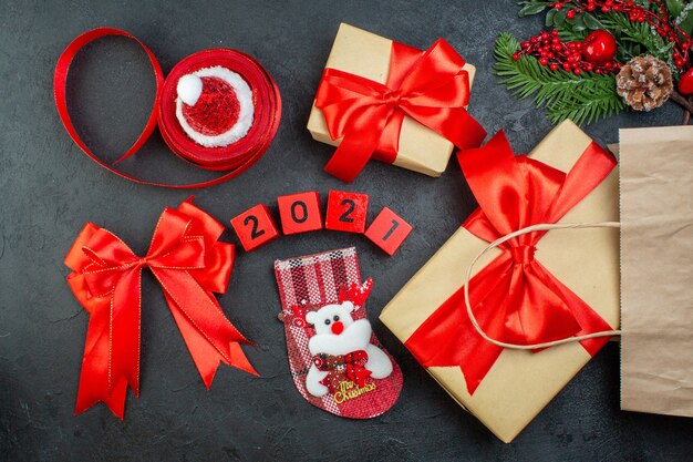 Vista anterior del estado de ánimo navideño con hermosos regalos ramas de abeto cono de coníferas cinta roja y números sombrero de santa claus calcetín de Navidad sobre fondo oscuro