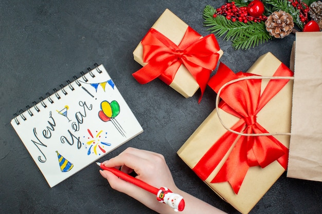 Vista anterior del estado de ánimo navideño con hermosos regalos y ramas de abeto cono de coníferas junto al cuaderno con dibujos de año nuevo sobre fondo oscuro