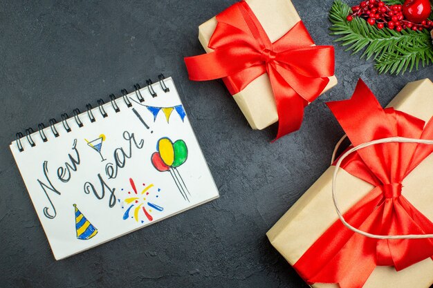 Vista anterior del estado de ánimo navideño con hermosos regalos y ramas de abeto cono de coníferas junto al cuaderno sobre fondo oscuro