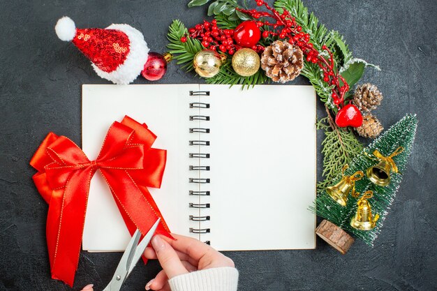 Vista anterior del estado de ánimo navideño con ramas de abeto, sombrero de santa claus, árbol de navidad, cinta roja en el portátil sobre fondo oscuro