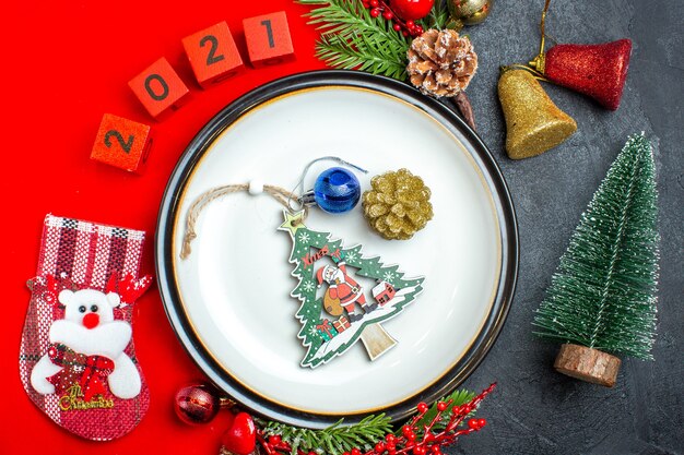 Vista anterior del fondo de año nuevo con accesorios de decoración de plato de cena ramas de abeto y números calcetín de navidad en una servilleta roja junto al árbol de navidad en una mesa negra