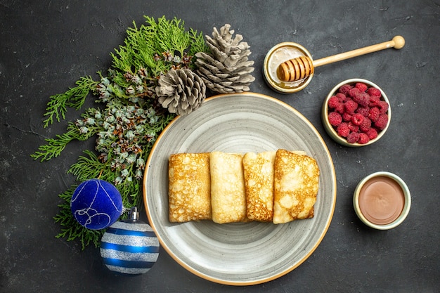 Vista anterior del fondo de la cena con deliciosos panqueques de miel y chocolate, frambuesa y cono de coníferas junto a accesorios de año nuevo sobre fondo negro