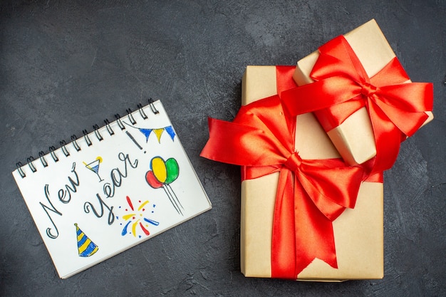 Vista anterior del fondo de Navidad con hermosos regalos con cinta en forma de arco y cuaderno con escritura de año nuevo sobre un fondo oscuro