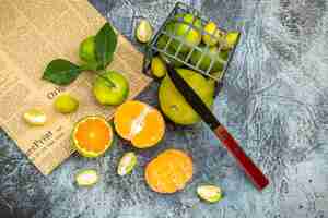 Foto gratuita vista anterior de frutas cítricas frescas con hojas caídas de una canasta negra cortada por la mitad y un cuchillo en un periódico sobre fondo gris