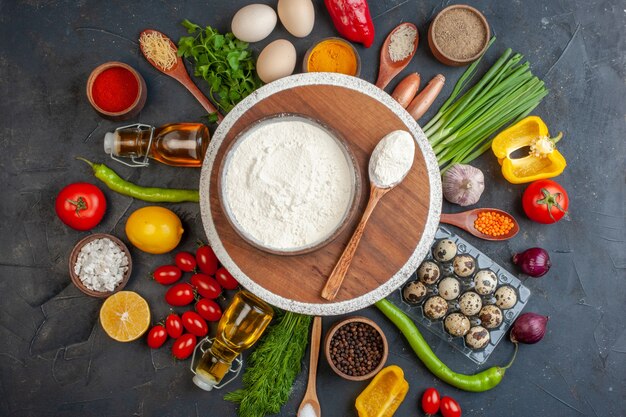 Vista anterior de harina en una olla marrón sobre tabla de cortar redonda entre verduras frescas botella de aceite de limón caído huevos diferentes especias en negro