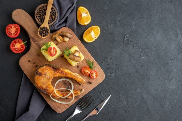 Vista anterior de harina de pescado frito con champiñones, verduras, queso sobre tabla de madera, rodajas de limón, pimienta sobre una toalla de color oscuro en el lado derecho sobre una superficie negra angustiada