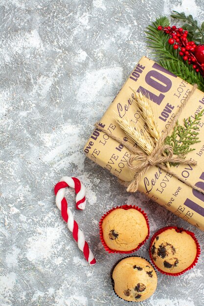 Vista anterior del hermoso regalo lleno de Navidad con inscripción de amor pequeños cupcakes dulces y accesorios de decoración de ramas de abeto cono de coníferas en la superficie del hielo