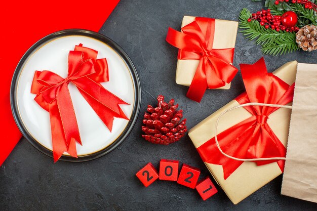 Vista anterior de hermosos regalos y cinta en forma de arco en un plato coníferas números de ramas de abeto de cono sobre una mesa oscura