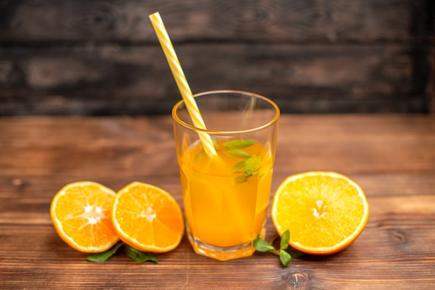 Vista anterior de jugo de naranja fresco en un vaso servido con un tubo de menta y limas de naranja sobre una mesa de madera