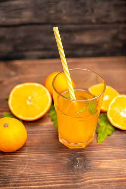 Vista anterior de jugo de naranja fresco en un vaso servido con un tubo de menta y naranjas enteras cortadas en una mesa de madera