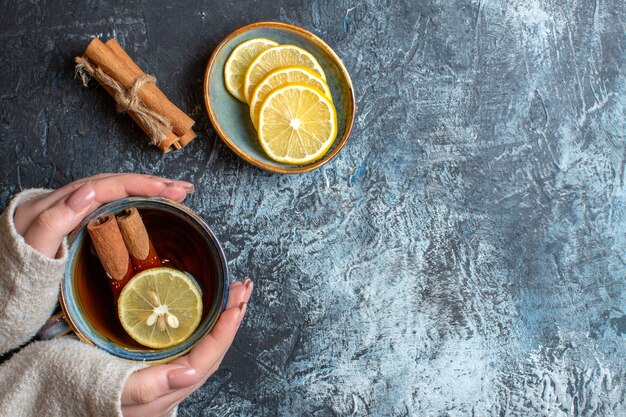 Vista anterior de limones frescos y mano sosteniendo una taza de té negro con canela sobre fondo oscuro