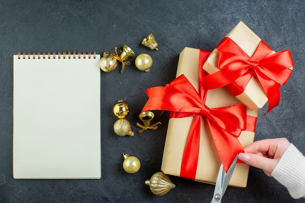 Vista anterior de la mano cortando la cinta roja en la caja de regalo y accesorios de decoración junto al cuaderno espiral sobre fondo oscuro