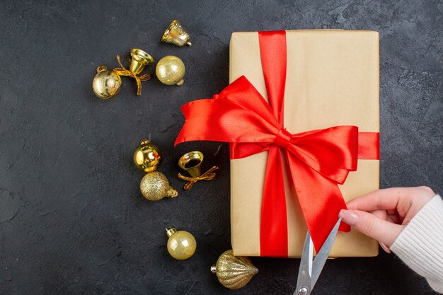 Vista anterior de la mano cortando la cinta roja en la caja de regalo y accesorios de decoración sobre fondo oscuro