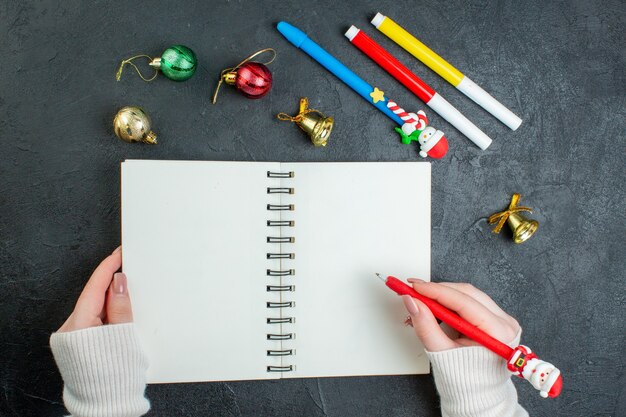 Vista anterior de la mano que sostiene un bolígrafo en el cuaderno espiral y accesorios de decoración sobre fondo negro