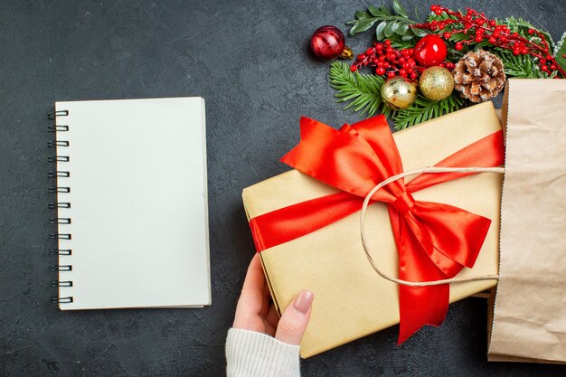 Vista anterior de la mano sacando una hermosa caja de regalo de una bolsa junto al portátil sobre fondo oscuro