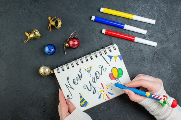 Vista anterior de la mano sosteniendo un bolígrafo en un cuaderno espiral con accesorios de decoración de dibujos y escritura de año nuevo sobre fondo negro