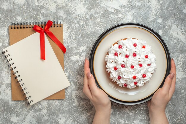 Vista anterior de la mano sosteniendo un plato con un delicioso pastel cremoso decorado con frutas junto a cuadernos sobre fondo de hielo