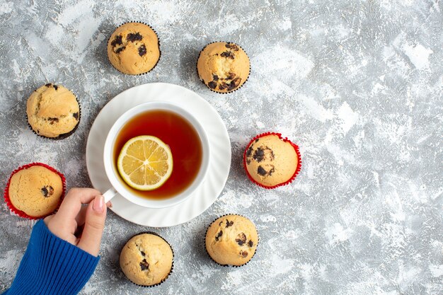 Vista anterior de la mano sosteniendo una taza de té y deliciosos cupcakes pequeños con chocolate sobre la superficie del hielo