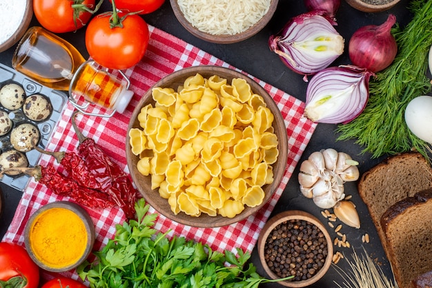 Vista anterior de pastas crudas en rojo toalla pelada verduras frescas rebanadas de pan botella de aceite caído un paquete verde sobre mesa negra