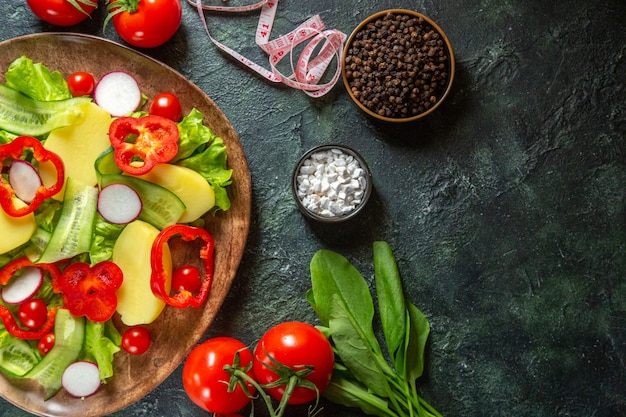 Vista anterior de patatas cortadas peladas frescas con pimiento rojo rábanos tomates verdes en una placa marrón y metros de especias en la superficie de colores mezcla verde negro