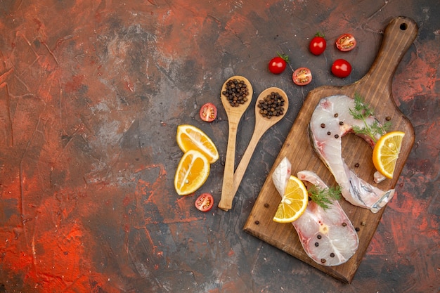 Vista anterior de pescado crudo y pimienta en la tabla de cortar de madera rodajas de limón tomates en la superficie de color de mezcla