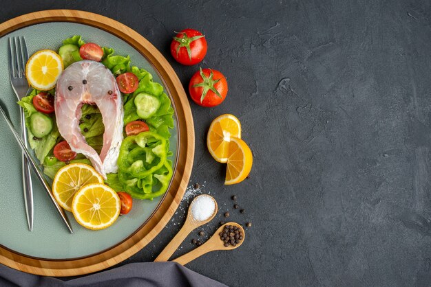 Vista anterior de pescado crudo y verduras frescas, rodajas de limón y cubiertos en una placa gris, especias, toalla de color oscuro en el lado izquierdo sobre una superficie negra