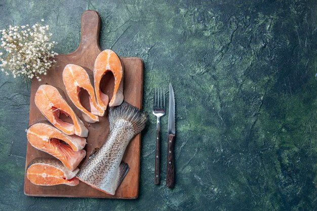 Vista anterior de pescados crudos frescos sobre una tabla de cortar de madera y un cuchillo en el lado derecho de la mesa de colores mezclados oscuros con espacio libre