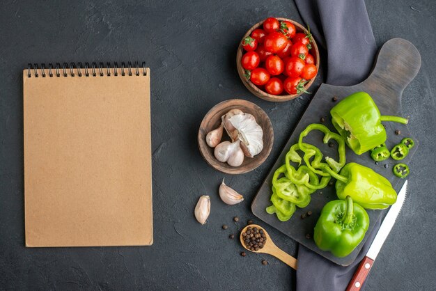 Vista anterior de pimientos verdes picados cortados enteros en la tabla de cortar de madera tomates en un tazón tomates ajos sobre una toalla de color oscuro sobre una superficie negra