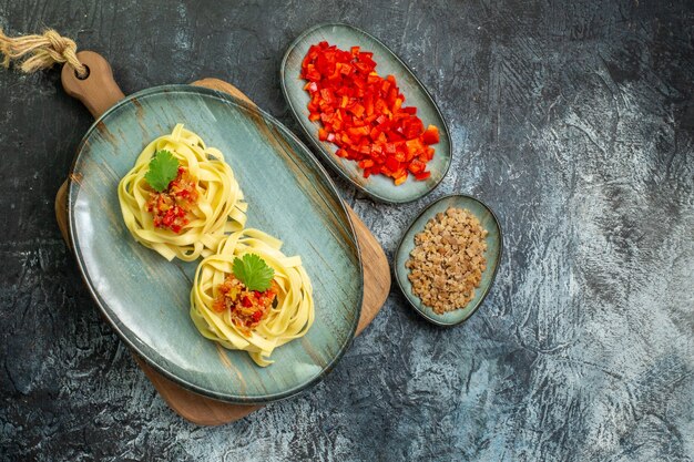 Vista anterior de una placa azul con una sabrosa comida de pasta servida con tomate y carne en la tabla de cortar junto a sus ingredientes en la mesa oscura