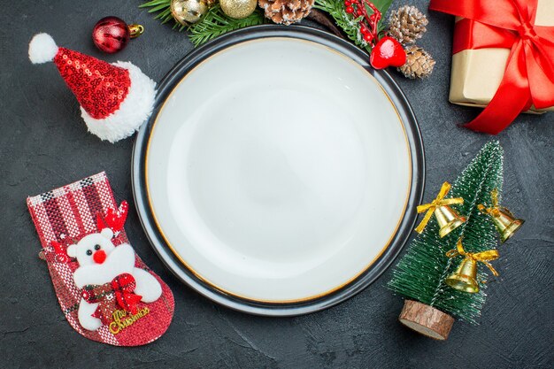 Vista anterior del plato de cena árbol de navidad ramas de abeto cono de coníferas caja de regalo sombrero de santa claus calcetín de navidad sobre fondo negro