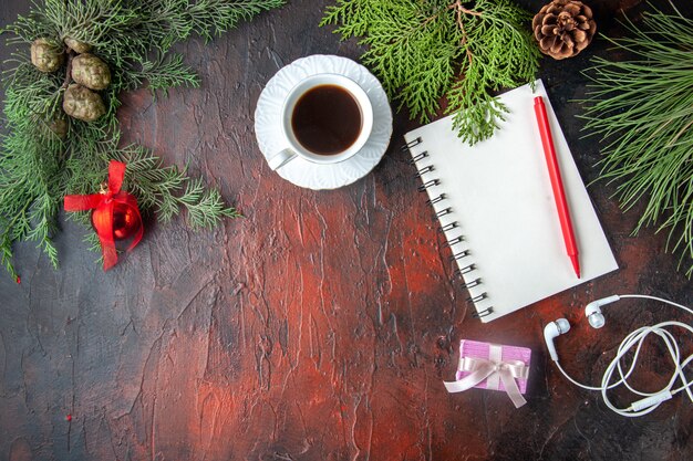 Vista anterior de ramas de abeto, una taza de té negro, accesorios de decoración, auriculares blancos y regalo junto al cuaderno con bolígrafo sobre fondo oscuro