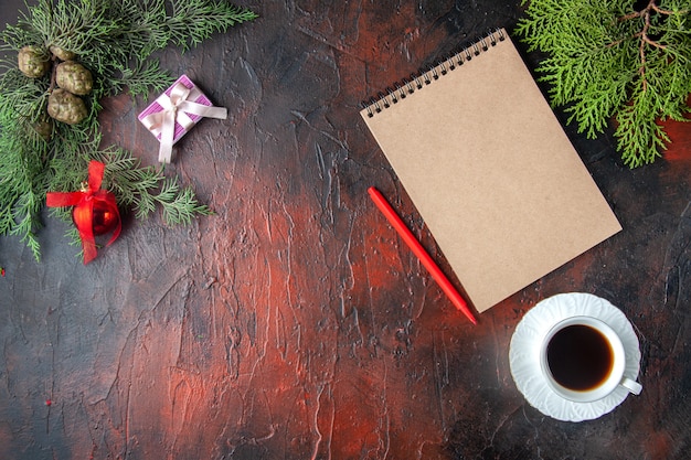 Vista anterior de ramas de abeto, una taza de té negro, accesorios de decoración y regalo junto al cuaderno con bolígrafo sobre fondo oscuro