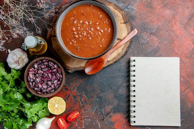 Vista anterior de la sopa de tomate en un recipiente azul en una bandeja de madera con cuchara, frijoles, botella de aceite, ajo y cuaderno en la tabla de colores mezclados