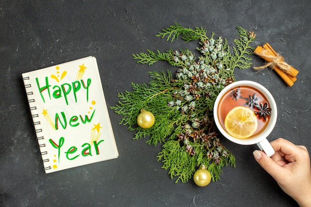 Vista anterior de una taza de té negro accesorios navideños y limas canela y cuaderno con inscripción feliz año nuevo sobre fondo negro