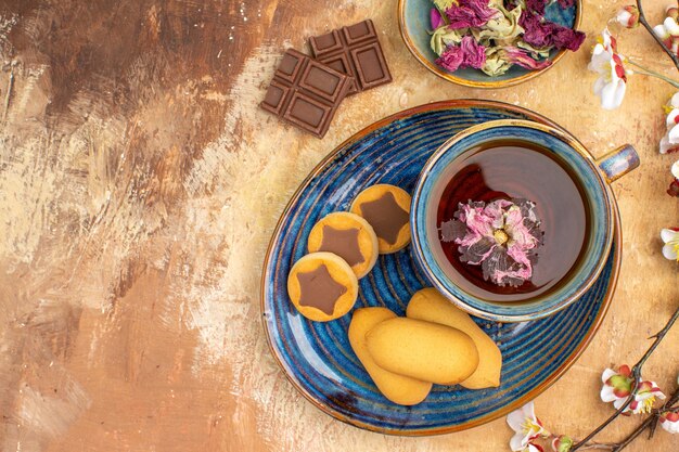 Vista anterior de varias galletas, una taza de té y barras de chocolate con flores en la tabla de colores mezclados