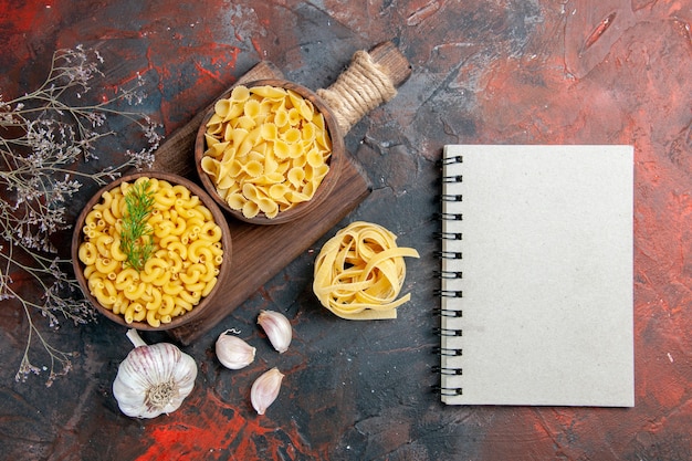 Vista anterior de varios tipos de pastas crudas en la tabla de cortar de madera, el ajo y el cuaderno en la tabla de colores mezclados