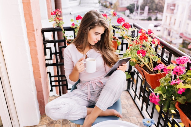 Vista desde arriba chica guapa en pijama desayunando en el balcón por la mañana en la ciudad. Ella sostiene una taza, leyendo en tableta.