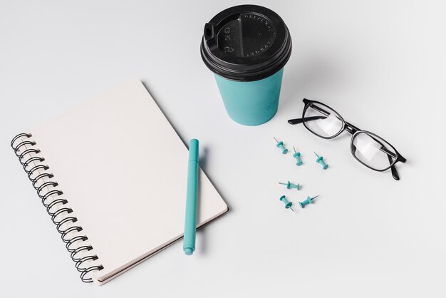 Una vista desde arriba del cuaderno espiral; bolígrafo; gafas; taza de café desechable; y chincheta sobre fondo blanco