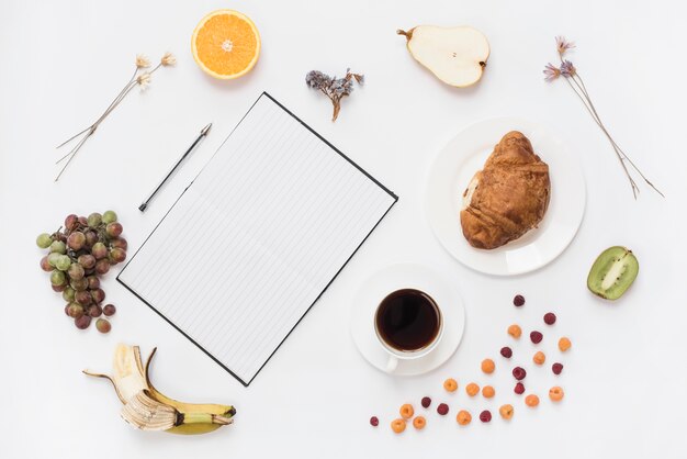 Una vista desde arriba del cuaderno; pluma con croissant y frutas saludables aisladas sobre fondo blanco