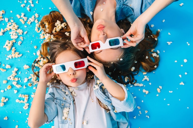 Vista desde arriba divertida madre e hija tendida en el piso, divirtiéndose con la cámara en palomitas de maíz aisladas sobre fondo azul. Familia de moda en ropa de jeans, con gafas 3D, expresando felicidad