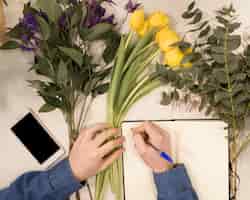 Foto gratuita una vista de arriba de una escritura masculina del florista en el diario con la pluma en la tabla con las flores