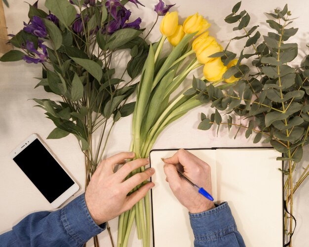 Una vista de arriba de una escritura masculina del florista en el diario con la pluma en la tabla con las flores