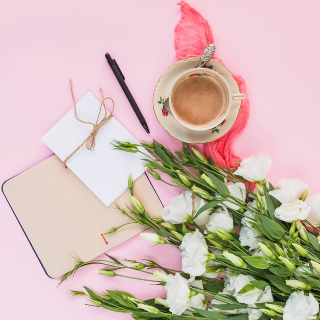 Una vista desde arriba de las flores de eustoma con diario; tarjeta; Taza de café y pluma sobre fondo rosa