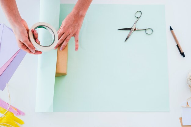 Una vista desde arriba de la mano de la mujer pegando el papel en la caja de regalo sobre el escritorio blanco