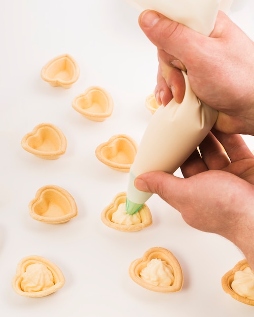 Vista de arriba de la mano que pone la crema en tartlet de la forma del corazón sobre el fondo blanco