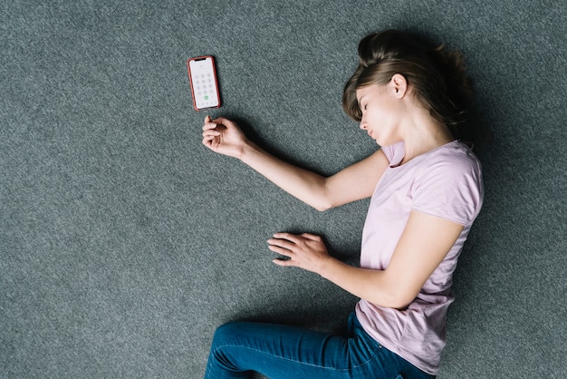 Vista de arriba de la mujer inconsciente que miente cerca del teléfono celular en la alfombra