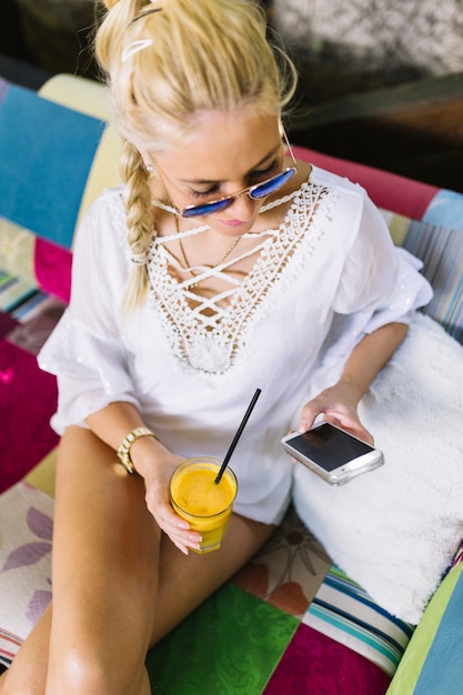 Foto gratuita una vista desde arriba de la mujer joven con estilo que usa el teléfono móvil que sostiene el vaso de jugo