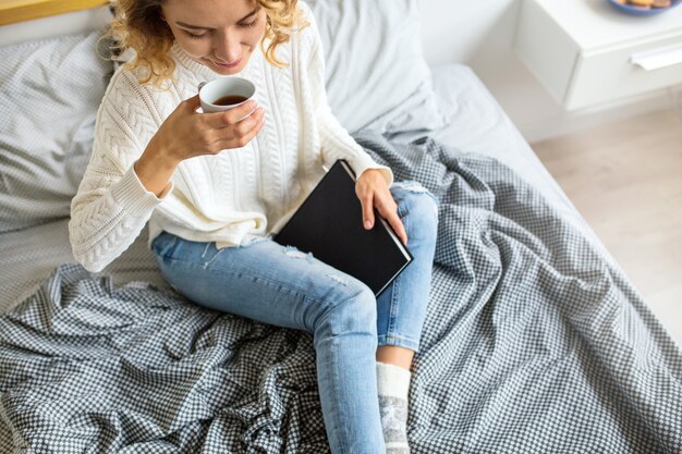 Vista desde arriba de la mujer sentada en la cama por la mañana, tomando café en la taza, sosteniendo el libro, vistiendo jeans